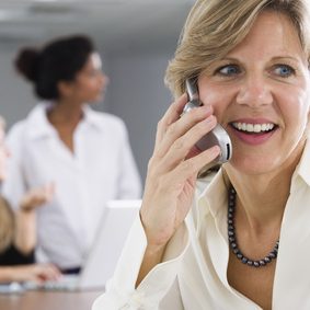 Frau spricht am Telefon, lächelt, während zwei Personen im Hintergrund diskutieren.