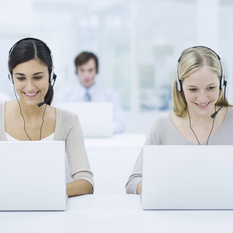 Zwei Frauen mit Headsets arbeiten an Laptops, ein Mann im Hintergrund.