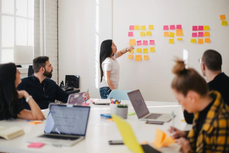 Teammeeting mit einer Präsentation an einer Wand aus Post-its. Laptops auf dem Tisch.