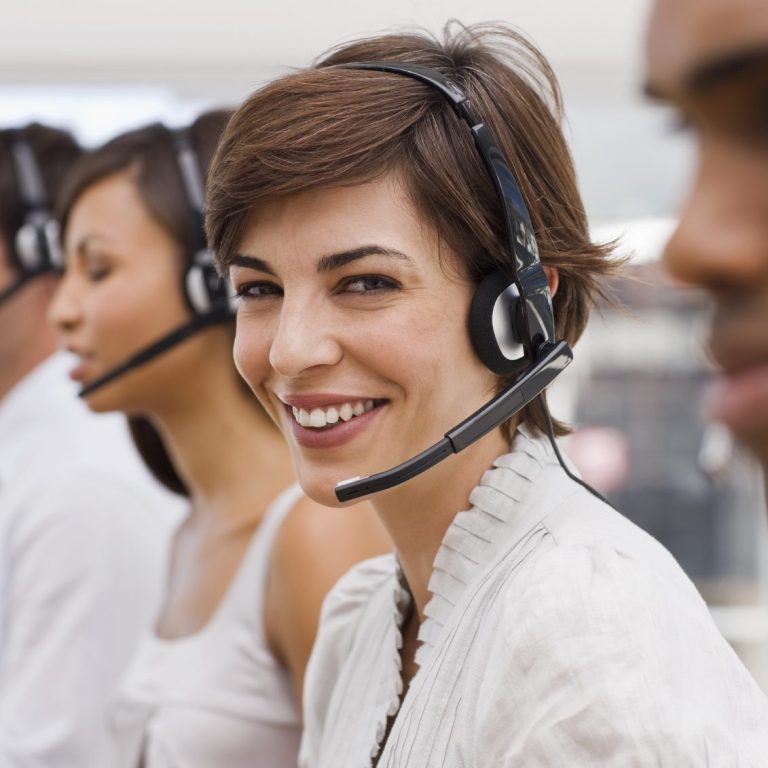 Lächelnde Frau mit Headset, umgeben von Kolleginnen und Kollegen in Callcenter-Umgebung.
